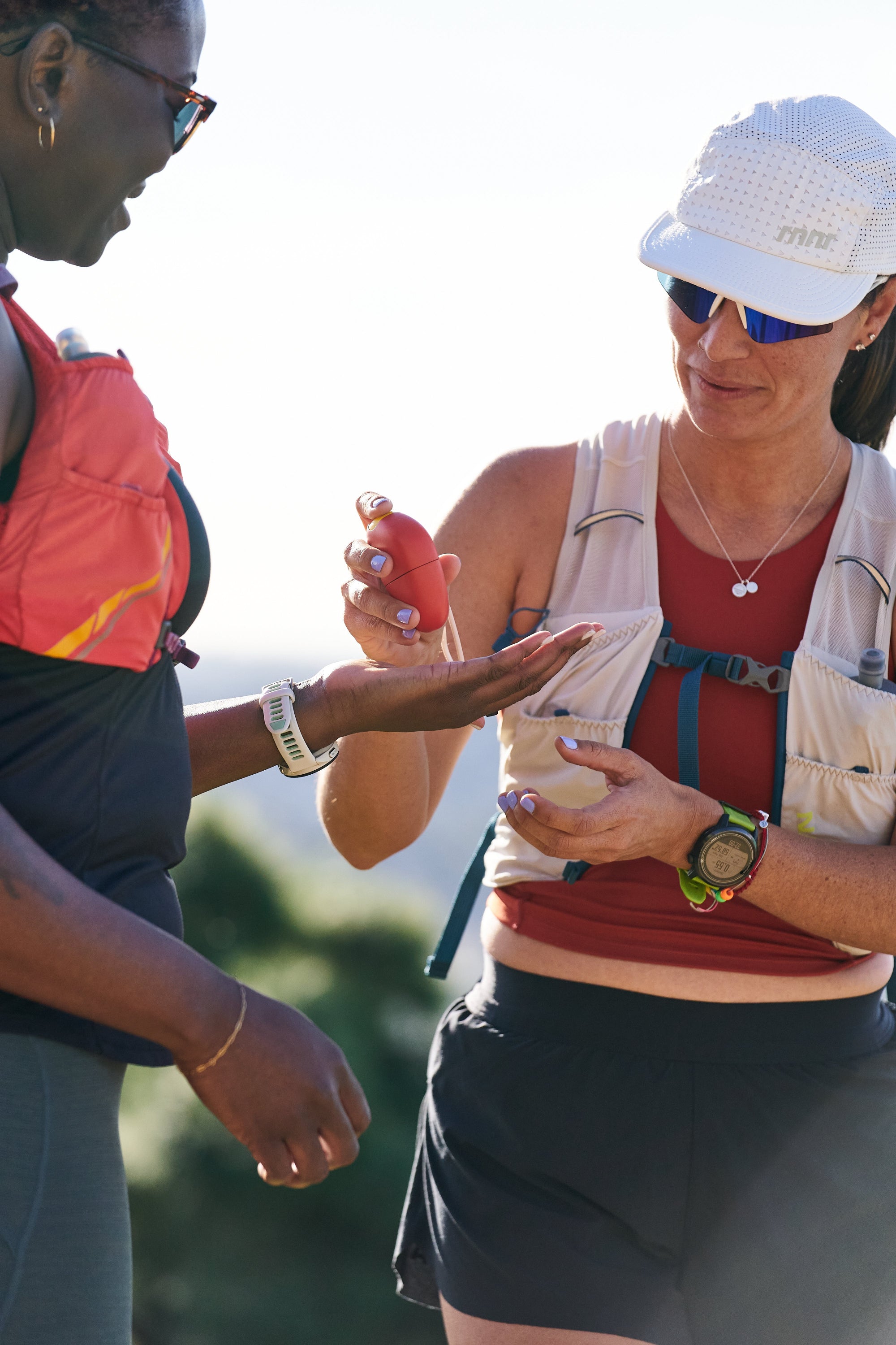 Two female hikers are on a trail, with one hiker handing sunscreen to the other.