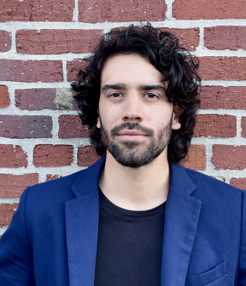 Camilo, dressed in a suit jacket, stands in front of a brick wall.